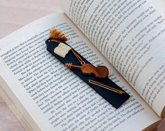 May include: A black leather bookmark with a wooden violin and bow, a book, and a tassel. The bookmark is decorated with a gold-colored chain.
