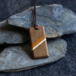 Könnte beinhalten: Ein rechteckiger Holzanhänger mit einer Basis aus dunkelbraunem Holz und einer diagonalen Einlage aus hellerem, beigefarbenem und cremefarbenem Holz. Der Anhänger hängt an einer dünnen, dunkelbraunen Kordel und hat eine glatte Oberfläche.