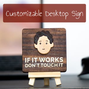 May include: A wooden desktop sign with a cartoon face and the text "IF IT WORKS DON'T TOUCH IT". The sign is on a small wooden easel.