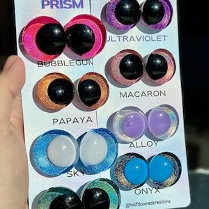 May include: Two white display cards labeled Half-Doomed Handcrafted Safety Eyes featuring pairs of round eyes in various colors including Bubblegum, Ultraviolet, Macaron, Papaya, Alloy, Sky, and Onyx. Each pair has a solid center and a glittery iris.
