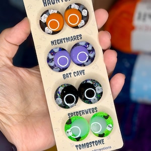 May include: A display card labeled Half-Doomed Handcrafted Safety Eyes featuring four pairs of circular eyes. The sets are titled Haunted with orange centers, Nightmares with purple centers, Bat Cave with black centers, and Spiderwebs with green centers.