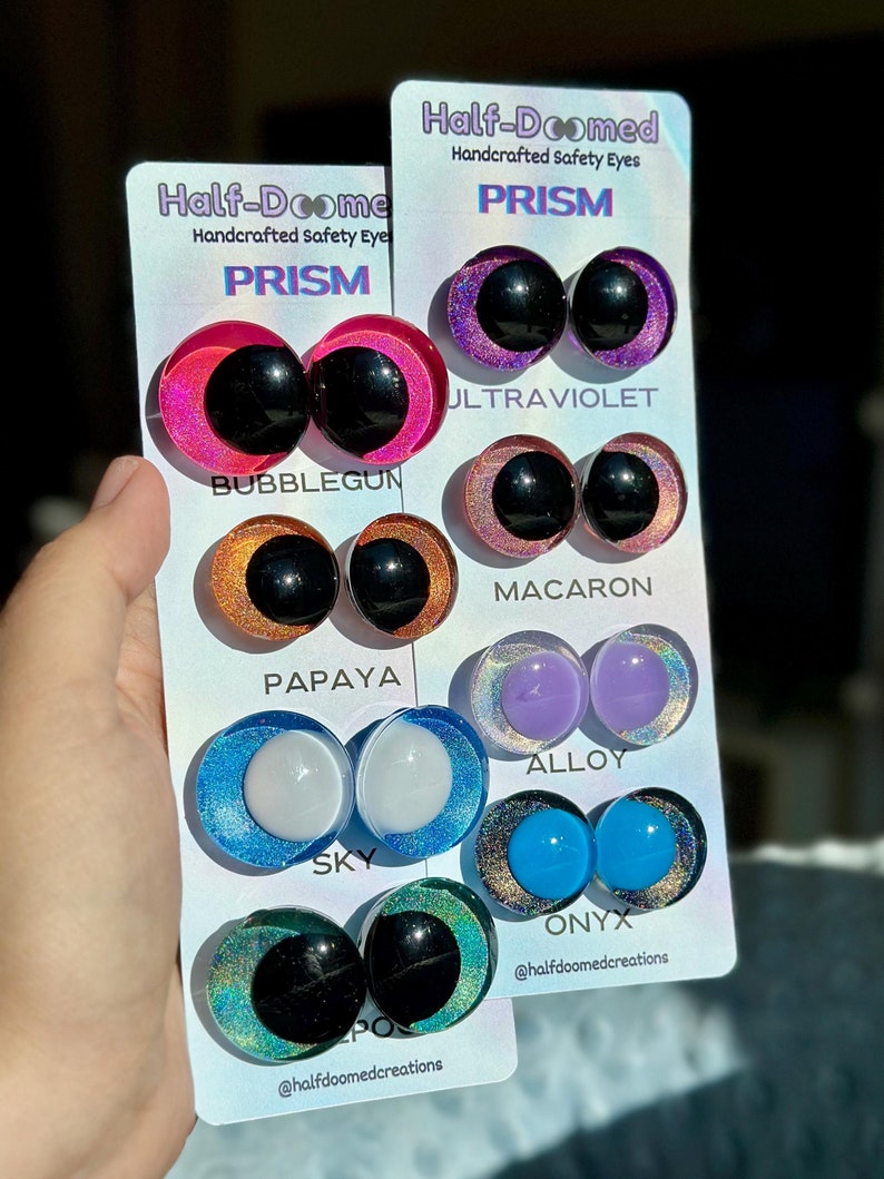 May include: Two white display cards labeled Half-Doomed Handcrafted Safety Eyes featuring pairs of round eyes in various colors including Bubblegum, Ultraviolet, Macaron, Papaya, Alloy, Sky, and Onyx. Each pair has a solid center and a glittery iris.
