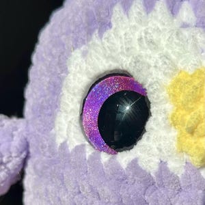 May include: A crocheted plush toy featuring a textured lavender yarn body with white and yellow accents. The eye is a large, glossy black circular piece surrounded by a crescent-shaped, glittery purple frame that catches the light.