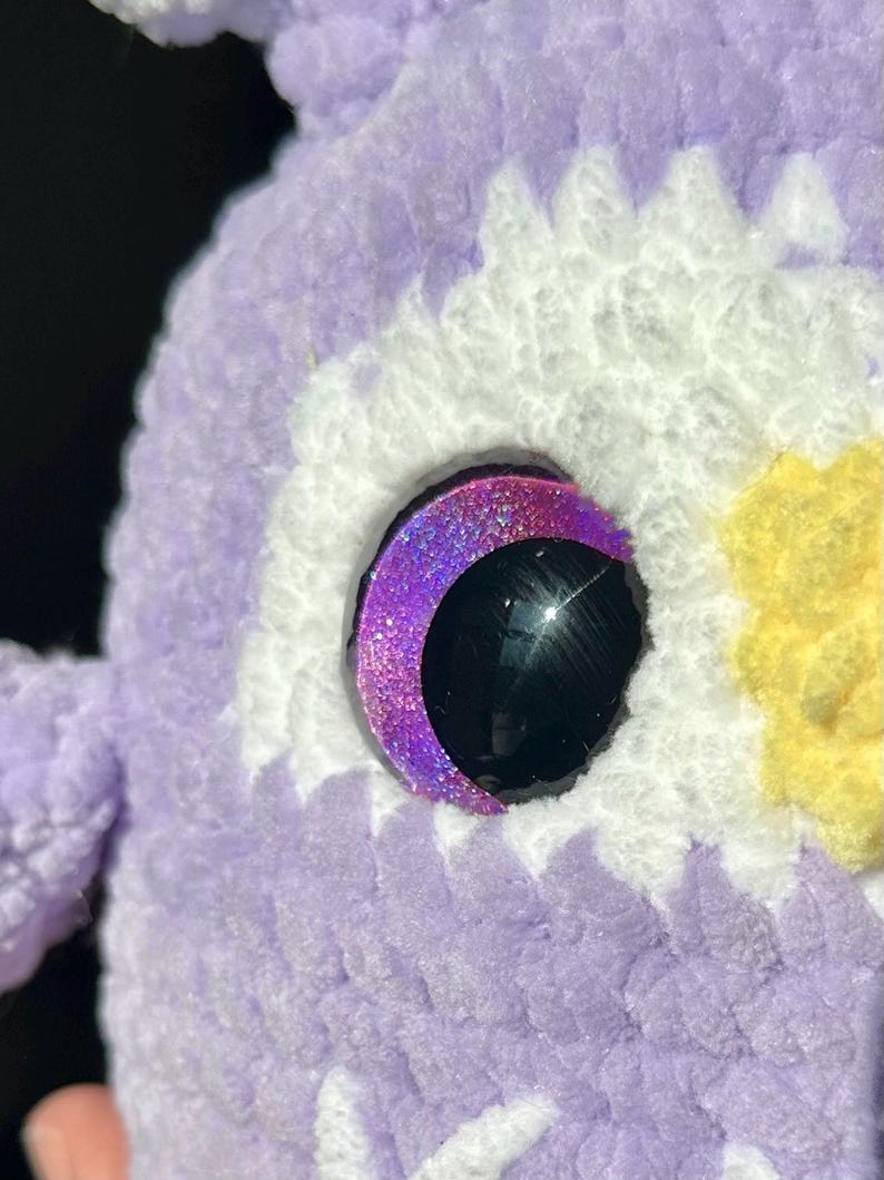May include: A crocheted plush toy featuring a textured lavender yarn body with white and yellow accents. The eye is a large, glossy black circular piece surrounded by a crescent-shaped, glittery purple frame that catches the light.