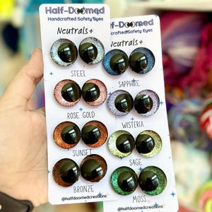 May include: A set of 10 pairs of glitter safety eyes for crafts. The eyes are arranged in two rows of five pairs. Each pair is labeled with a color name: Steel, Sapphire, Rose Gold, Sunset, Bronze, Wisteria, Sage, Moss. The eyes are all black with a glitter finish.