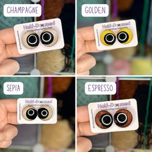 May include: Four sets of handcrafted safety eyes on white display cards labeled Half-Doomed. The colors include champagne, golden, sepia, and espresso. Each pair features a round, glittery iris with a large black pupil and a white ring accent.
