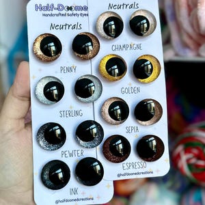 May include: Two white display cards labeled Half-Doomed Handcrafted Safety Eyes Neutrals featuring pairs of round, glittery safety eyes in colors including penny, sterling, pewter, ink, champagne, golden, sepia, and espresso with black pupils.