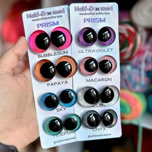 May include: Two white display cards labeled Half-Doomed Handcrafted Safety Eyes Prism featuring eight pairs of round, glittery safety eyes in colors Bubblegum, Ultraviolet, Papaya, Macaron, Sky, Alloy, Tidepool, and Onyx with black pupils.