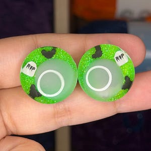 以下が含まれることがあります： 鮮やかなネオングリーンのグリッターベースに、黒いコウモリのシルエットと黒い文字でRIPと書かれた小さな白い墓石のグラフィックが描かれた、円形のイヤープラグのペア。各プラグの表面には中央に白いリングのデザインがあります。