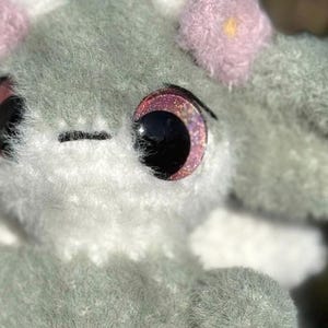 May include: A plush toy crafted from fuzzy sage green and white yarn. The creature features large, round, dark eyes with pink glittery irises, a small black stitched mouth, and soft pink flower-shaped accents attached to the top of its head.