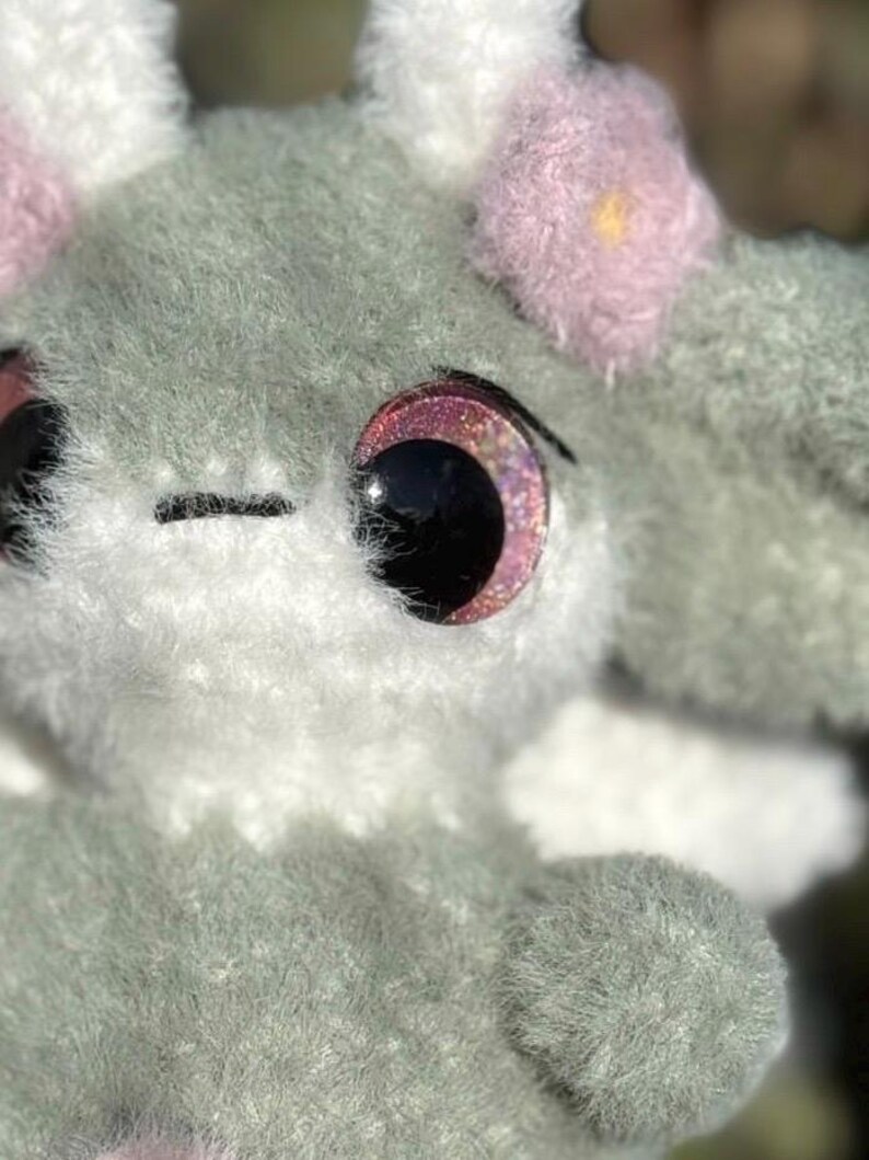 May include: A plush toy crafted from fuzzy sage green and white yarn. The creature features large, round, dark eyes with pink glittery irises, a small black stitched mouth, and soft pink flower-shaped accents attached to the top of its head.