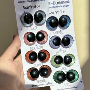 May include: Two white display cards labeled Half-Doomed Handcrafted Safety Eyes Neutrals+ featuring eight pairs of glittery safety eyes in steel, sapphire, rose gold, wisteria, sunset, sage, bronze, and moss colors with black pupils.