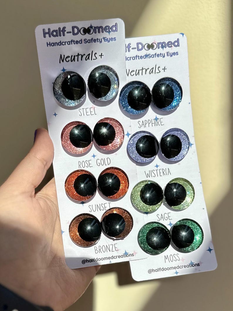 May include: Two white display cards labeled Half-Doomed Handcrafted Safety Eyes Neutrals+ featuring eight pairs of glittery safety eyes in steel, sapphire, rose gold, wisteria, sunset, sage, bronze, and moss colors with black pupils.