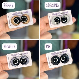 May include: Four sets of circular safety eyes on white display cards labeled Penny, Sterling, Pewter, and Ink. Each pair features a black center with a white ring and a glittery outer edge in copper, silver, dark gray, or black. Text reads Half-Doomed.