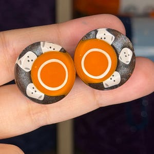 以下が含まれることがあります： 暗いグリッター調の背景に、白い縁取りの鮮やかなオレンジ色の円が中央に配置された円形のイヤープラグのペア。オレンジ色の中心の周りには、黒い点の目と口を持つ小さな白い幽霊のような図形が描かれています。