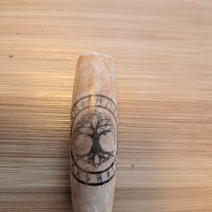 Op de afbeelding: Een houten cilinder met een levensboomontwerp en runen rond de buitenkant. De cilinder is lichtbruin en heeft een glad oppervlak.