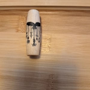 Op de afbeelding: Een houten pen met zwarte inktmarkeringen. De markeringen zijn abstract en lijken op een gestileerde vogel met hangende ornamenten.