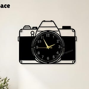 Puede incluir: Un reloj de pared en blanco y negro con forma de cámara. La esfera del reloj es redonda con manecillas doradas y números negros. El texto "enchantingspace" está en la esquina superior izquierda.