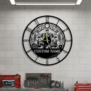 May include: Black metal wall clock with a mechanic theme. The clock features a silhouette of a mechanic, a car, and tools. The text "CUSTOM NAME" is displayed on the clock face. The clock is hanging on a white brick wall in a garage setting.