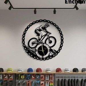 Pode incluir: Um relógio de parede de metal preto com um ciclista em uma bicicleta de montanha, emoldurado por uma corrente. O mostrador do relógio mostra uma paisagem de montanha com ponteiros indicando a hora. A palavra "Enchantingspace" está no topo.