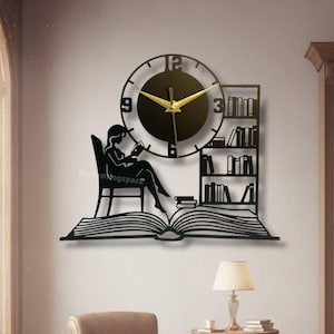 Könnte beinhalten: Schwarze Metall-Wanduhr mit einer Silhouette einer lesenden Person, einem Bücherregal und einem offenen Buch. Die Uhr hat goldene Zeiger und ein schwarzes Zifferblatt. Der Text "Enchantingspace" befindet sich am unteren Bildrand.