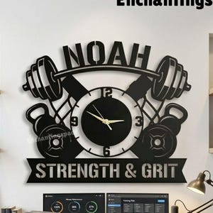 May include: Black metal wall clock with the name "NOAH" above a barbell and kettlebell design. The clock face is surrounded by weights and the words "STRENGTH & GRIT" are below. The background includes weights, a desk with computers, and the text "Enchantingspace".