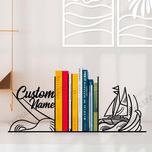 Puede incluir: Sujeta libros de metal negro con forma de velero sobre olas con el texto "Custom Name" en el lado izquierdo. Los sujeta libros sostienen una fila de libros coloridos.