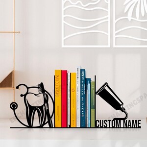 Könnte beinhalten: Schwarze Metall-Buchstützen in Form von Zahnarztinstrumenten, darunter eine Zahnbürste, ein Zahn und ein Stethoskop. Die Buchstützen halten eine Reihe bunter Bücher. Der Text "CUSTOM NAME" ist auf der Basis der Buchstützen aufgedruckt.