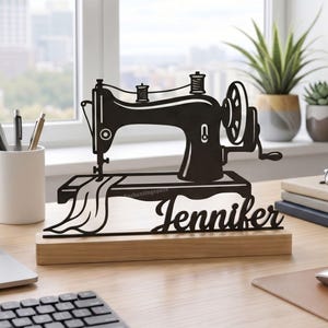 May include: A black silhouette of a vintage sewing machine on a wooden base. The name "Jennifer" is displayed below the machine. The text "Enchantingspace" is visible at the top. The desk also has a laptop, keyboard, and other office supplies.