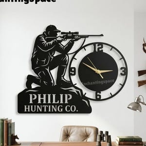 May include: Black metal wall clock featuring a hunter aiming a rifle, with the text "PHILIP HUNTING CO." below. The clock has gold hands and the text "Enchantingspace". Decorative hunting-themed wall art.