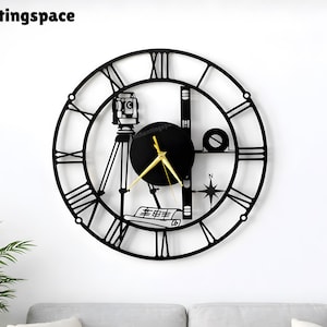 Op de afbeelding: Zwarte metalen wandklok met Romeinse cijfers en gouden wijzers. De klok heeft landmeetkundige gereedschappen, waaronder een camera op een statief en een kompas. De klok is gemonteerd op een witte muur boven een grijze bank.