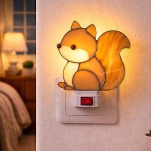 Puede incluir: Una luz de noche de vidriera con forma de ardilla, con tonos naranja y marrón. La luz de noche está enchufada a un enchufe blanco con un interruptor de encendido/apagado rojo. El texto "Enchantingspace" es visible en la parte inferior.