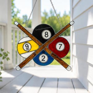 Può includere: Acchiappasole in vetro colorato con palle da biliardo e stecche. Il design presenta le palle 8, 7, 9 e 2, con due stecche incrociate. L'acchiappasole è sospeso da una catena. Il testo "Enchantingspace" è visibile.