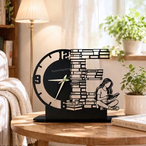 Può includere: Un orologio da scrivania nero con un quadrante circolare e un design decorativo di libri e una persona che legge. Il quadrante ha lancette bianche e il testo "Enchantingspace". L'orologio è su un tavolo di legno.