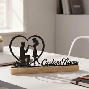 May include: A black silhouette of a marriage proposal scene, featuring a kneeling person offering a ring to another person inside a heart-shaped frame. The design sits on a wooden base with the text "Custom Name". The brand name "Enchantingspace" is visible.