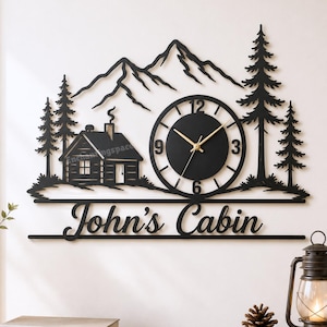 Könnte beinhalten: Schwarze Metall-Wanduhr mit einem Design aus Hütte, Bergen und Bäumen. Das Zifferblatt ist rund mit goldenen Zeigern und dem Text "John's Cabin" darunter. Ein dekoratives Stück für ein rustikales Zuhause.