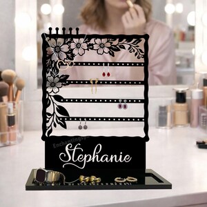 May include: A black acrylic jewelry organizer with a floral design and the name "Stephanie" in white script. The organizer has multiple horizontal bars for hanging earrings and a base for storing rings and bracelets. The background shows a person applying makeup.