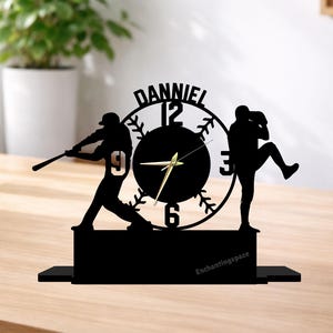 Peut inclure: Horloge noire sur le thème du baseball avec une silhouette de frappeur et de lanceur. Le cadran de l'horloge affiche les chiffres 12, 3, 6 et 9, avec le nom "DANIEL" au-dessus. L'horloge est sur une surface en bois, avec le texte "Enchantingspace" en bas.