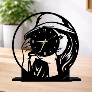 May include: Black and white decorative clock featuring a woman wearing a hat, holding the clock face. The clock has gold hands and a black face. The base of the clock is black, and the text "Enchantingspace" is at the bottom.