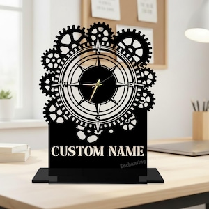 May include: Black desk clock with a compass design, surrounded by gears. The clock face features a compass rose and hands. The base of the clock has the words "CUSTOM NAME" printed on it. The clock is on a desk with a lamp and a corkboard in the background.