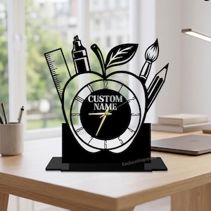 May include: Black desk clock in the shape of an apple, with a clock face and school supplies. The clock has the words "CUSTOM NAME" on the face. The clock is on a black base and the brand name "Enchantingspace" is visible.