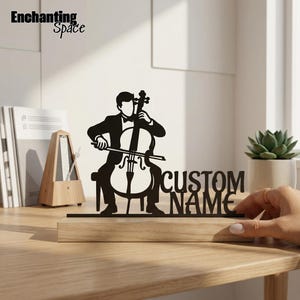 Könnte beinhalten: Eine schwarze Metallsilhouette eines Musikers, der Cello spielt, montiert auf einem hellen Holzsockel. Das Design enthält den Text CUSTOM NAME, der in die Struktur integriert ist. Das Objekt steht auf einer Holzfläche neben einem Metronom.