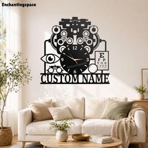 May include: A black wall clock featuring an optometrist-themed design with a phoropter, an eye icon, a Snellen eye chart, and a circular clock face with gold hands. The base includes the text CUSTOM NAME in a bold, sans-serif capital font.