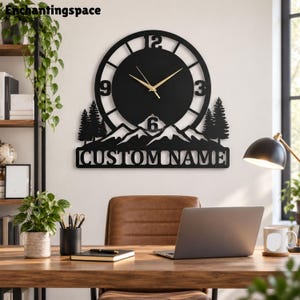 May include: A black wall clock featuring a mountain range silhouette with pine trees and the text CUSTOM NAME. The clock face has a circular design with large numerals 3, 6, 9, and 12, complemented by thin gold-colored clock hands.