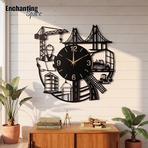 May include: A black wall clock featuring a construction theme with a crane, a bridge, a building, and heavy machinery silhouettes. The circular clock face has gold hands and numbers 3, 6, 9, and 12. The text Enchanting Space is visible.