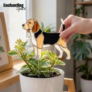Puede incluir: Un adorno de vitral para plantas con forma de perro beagle, compuesto por segmentos de vidrio blanco, beige y negro unidos por líneas de soldadura metálica. La figura está montada sobre una varilla de metal. El texto Enchanting Space es visible.