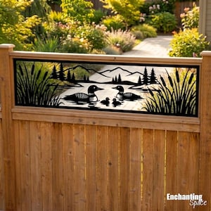 May include: A black metal fence insert featuring a laser-cut silhouette of two adult loons and two chicks swimming on a lake, surrounded by cattails and pine trees with a mountain range in the background, mounted on a wooden fence panel.
