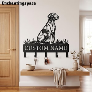 Puede incluir: Un soporte para llaves de pared de metal negro con la silueta de un perro sentado sobre un parche de hierba. El diseño incluye un panel rectangular con el texto CUSTOM NAME y cinco ganchos orientados hacia abajo para colgar llaves.