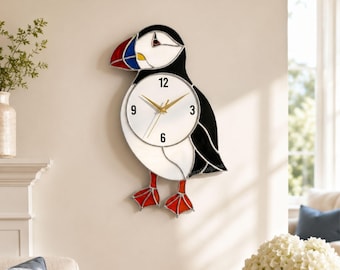 May include: A wall clock shaped like a puffin bird, featuring a white face with black numerals at 12, 3, 6, and 9. The clock has a stained glass style design with black, white, red, and blue sections, gold-colored hands, and bright orange feet.