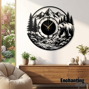 Puede incluir: Un reloj de pared circular de metal negro con un diseño cortado a láser de una cordillera, pinos, un oso de pie en una orilla rocosa y un pez saltando fuera del agua. La esfera del reloj tiene manecillas doradas y números negros.
